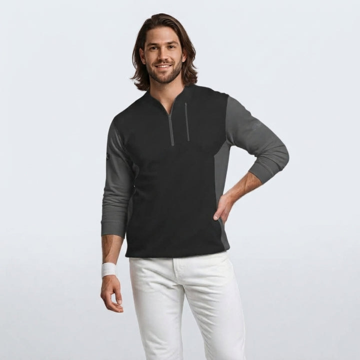 A man modeling a black and gray color-blocked long-sleeve quarter-zip pullover, paired with white pants, against a light gray studio background.